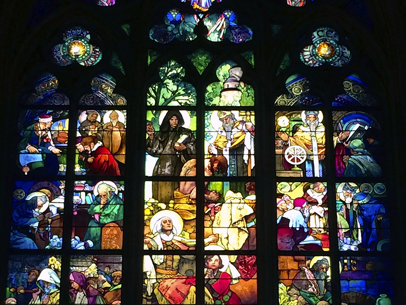 Backlit stained glass windows depicting many figures performing various actions, like kneeling, praying, writing.