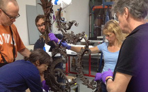 five gloved adults standing around a large ornate piece of metal, apparently very old and protected with tissue and styrofoam.