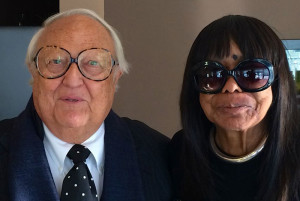A white-haired gentleman in oversized tortoiseshell glasses and polka-dotted tie. To his left is a brown-haired woman sporting oversized sunglasses and eyebrow-length bangs.