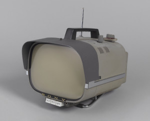 Portable television with square screen set in long rectangular gray metal housing with rounded edges; moveable visor at front; black plastic handle at top, retractable antenna at top left rear, station dial and volume control dial at top right rear; three square white control buttons on bottom front under the name SONY; metal base.