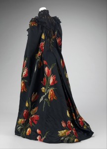 Photo of a mannequin against a photo shoot backdrop wearing a long black cloak with red and yellow sprays of tulips scattered around. The mannequin faces away from the camera showing the back of the cloak, which reaches down to the floor.