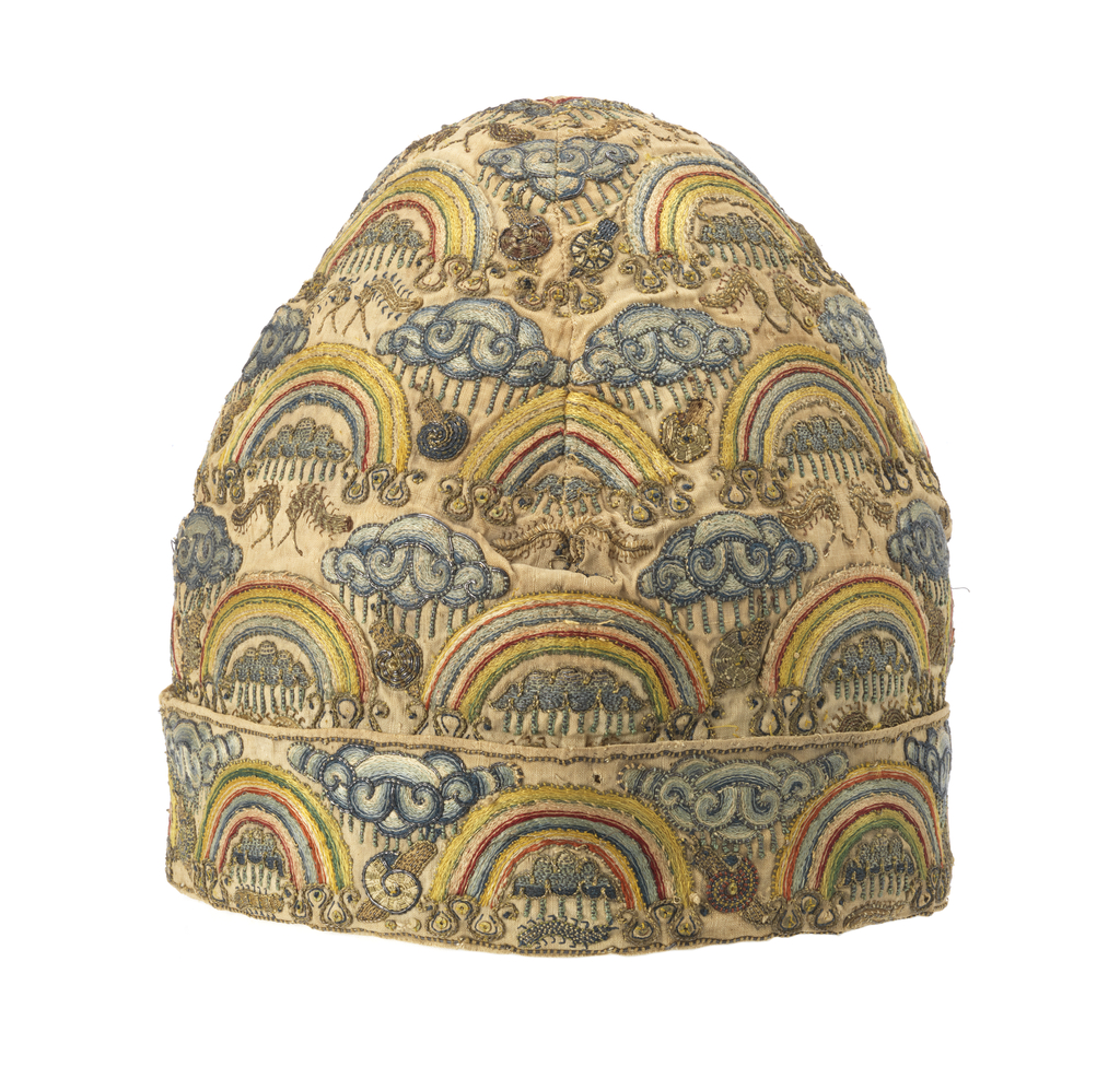 A man's conical cap with turned up cuff of off-white linen, embroidered in a pattern of rainbows hovering over clouds with rain falling on snails and caterpillars, in blue, green, yellow, red and pink silk and silver metallic threads.