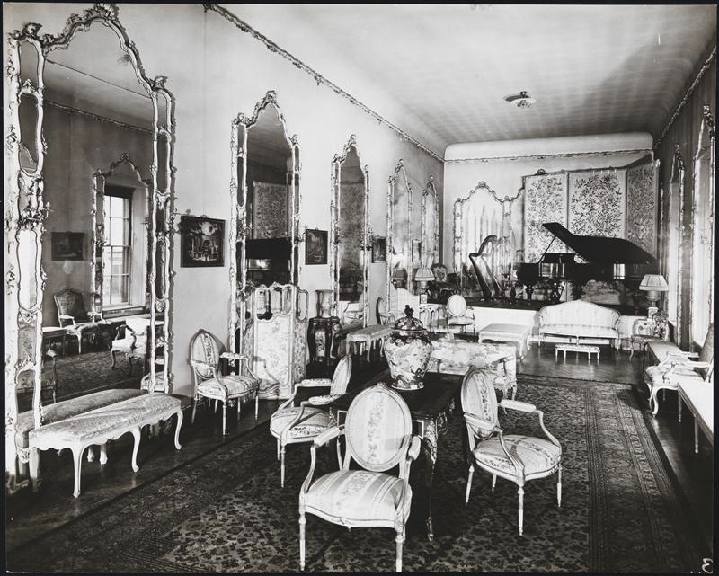 Image of an ornate rectangular room with 6 large gilded mirrors on one wall, about 6 cushioned chairs, several tables, benches and a black grand piano in back of room.