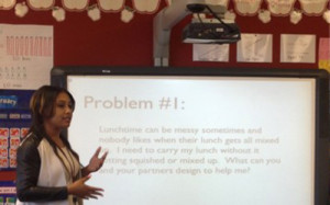 Woman gesturing in front of a classroom. Behind her a projected slide that says "Problem #1:" with more text below.