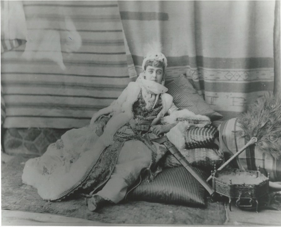 Woman laying down on a pile of pillows in lots of layered, drapey clothing. Sword on the ground to her left and a peacock feather fan beside it.