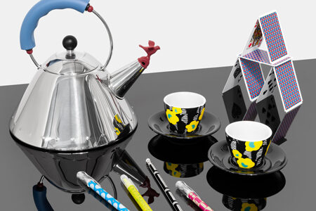 A kettle, espresso cups, pens, and a house of cards grouped together on a reflective black surface against an off-white background