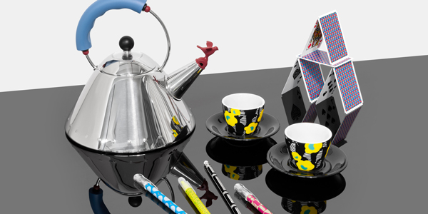 A kettle, espresso cups, pens, and a house of cards grouped together on a reflective black surface against an off-white background