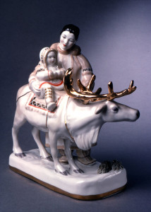 Woman stands next to a large reindeer, holding a young child sitting on the reindeer; both dressed in fur-lined parkas and boots; rectangular base.