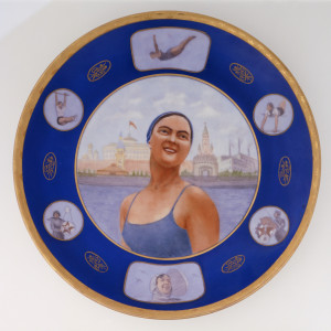 Large, circular form; in center, image of smiling woman dressed in bathing suit and bathing cap, standing before skyline of Moscow; blue border with six reserves showing women athletes; between each reserve an oval medalion with "USSR" (in Russian) and floral motif; gilded border with burnished star-like pattern.