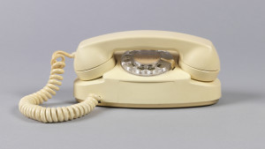 White telephone with oval body, handset cradled horizontally across top above clear plastic rotary dial; white coiled cord on left connecting handset to body.
