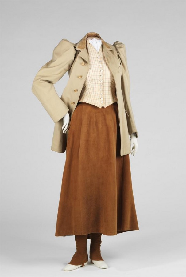 Mannequin in long tan dress, matching tan stockings which cover most of the white shoes below, and a poofy-shouldered tan jacket over a checkered ivory vest. Mannequin has one hand on hip.