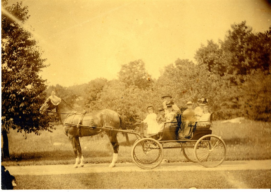 sepia toned photograph of a horse drawn carriage filled with children in white dresses and a few adults. blurry old picture. the horse wears a lacey hat on its head and ears.