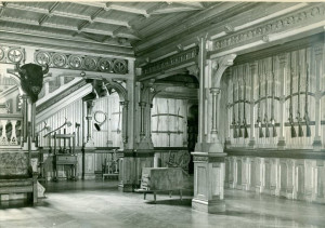 black and white photo of a hallway with detailed wood ornamentation covering every wall and archway, lots of rifles hanging on the wall, and a buffalo head on the wall too.
