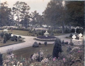 a formal garden with orderly quadrants divided by paved paths. A round circle of plantings and stone statue, appears to be a big vase, in the center.