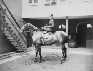 black and white photo of a woman on a horse in full riding getup, including whip and hat. Background is a spacious and clean stable interior.