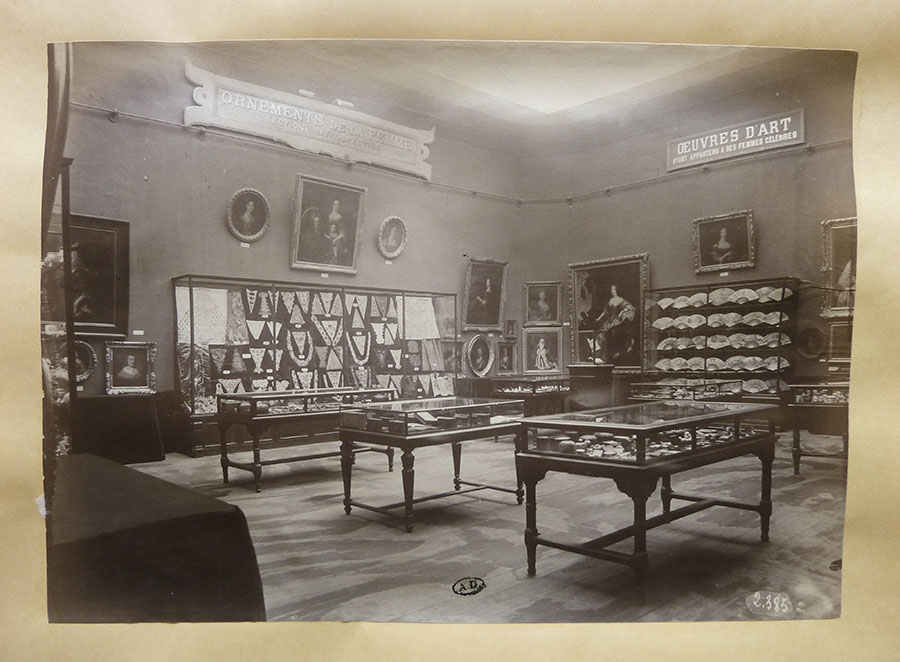 Black and white photo of a room with tables topped with fitted glass cases, displaying objects inside. Paintings line the walls and more objects in a class case on the wall.