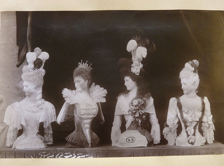 Four mannequins, from the waist up, placed in a row on a table, looking sharply over their right shoulders, festooned with feathers, elaborate lace, flowers and lofty hairdos.