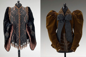 Two headless dressforms side by side showing garments. On the left, a heavy velvet top with draping black velvet sleeves, very loose around the hands in a medieval-looking way. Large leaf pattern on the front chest and a row of tassle-like dangling beads down the center line of the top and bottom waistline area. On the right, brown velvet coat with puffy balloon shoulders and large green bow at the neck. Glittery designs around the belly area, making a pattern of horizontal arches.