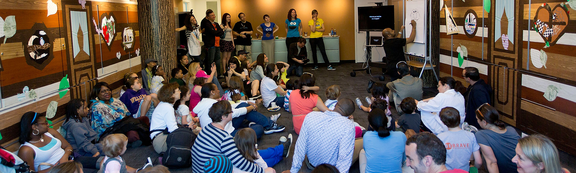 About 20 children and their families sitting on the floor, watching a man draw a sesame street character on an easel.