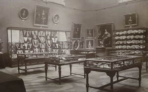 sepia toned photo of a room with three glass cases full of objects on view and paintings and jewelry displayed in frames on the walls.