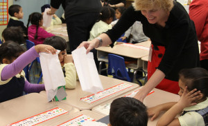 A female teacher holds a white paper bag and leans over a classroom table to show a group of four children a class project.