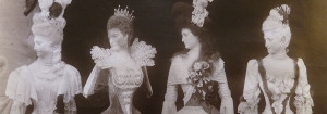 Four mannequins, from the waist up, placed in a row on a table, looking sharply over their right shoulders, festooned with feathers, elaborate lace, flowers and lofty hairdos.