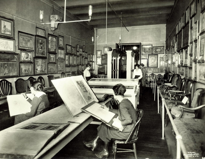 Photo of a large room. Walls covered in framed works. Women in tall leather boots with long dresses and long hair sit hunched over sketchboards, appearing to copy other works set in front of them on the table.