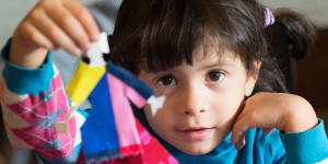 Young girl, about 4, holding a craft project up, something made of colorful paper, and gazing at the camera.