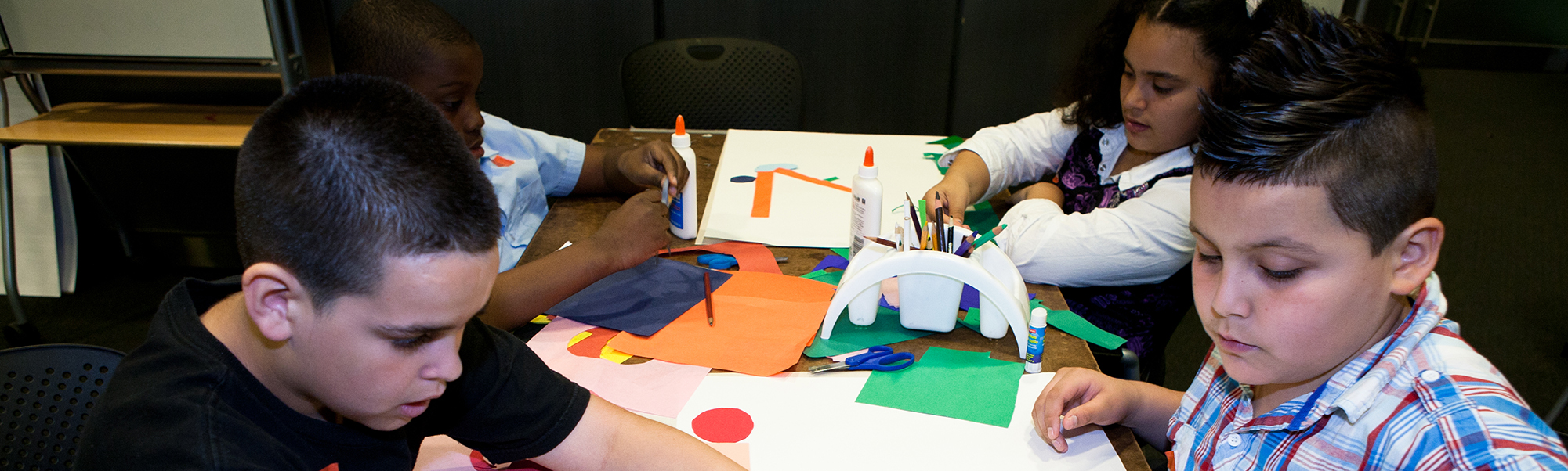 four elementary school children seated at a table, gluing strips of colored paper onto posterboard.