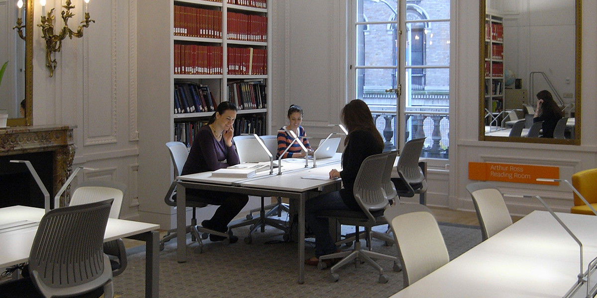 A clean white room with ornate gold sconces, a large mirror and fireplace, new white desks and rolling chairs, bookshelf and large windows. Three women sit at one of the tables, with books out, chatting.