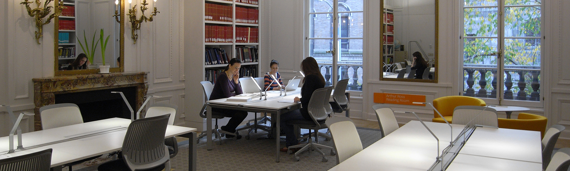 A clean white room with ornate gold sconces, a large mirror and fireplace, new white desks and rolling chairs, bookshelf and large windows. Three women sit at one of the tables, with books out, chatting.