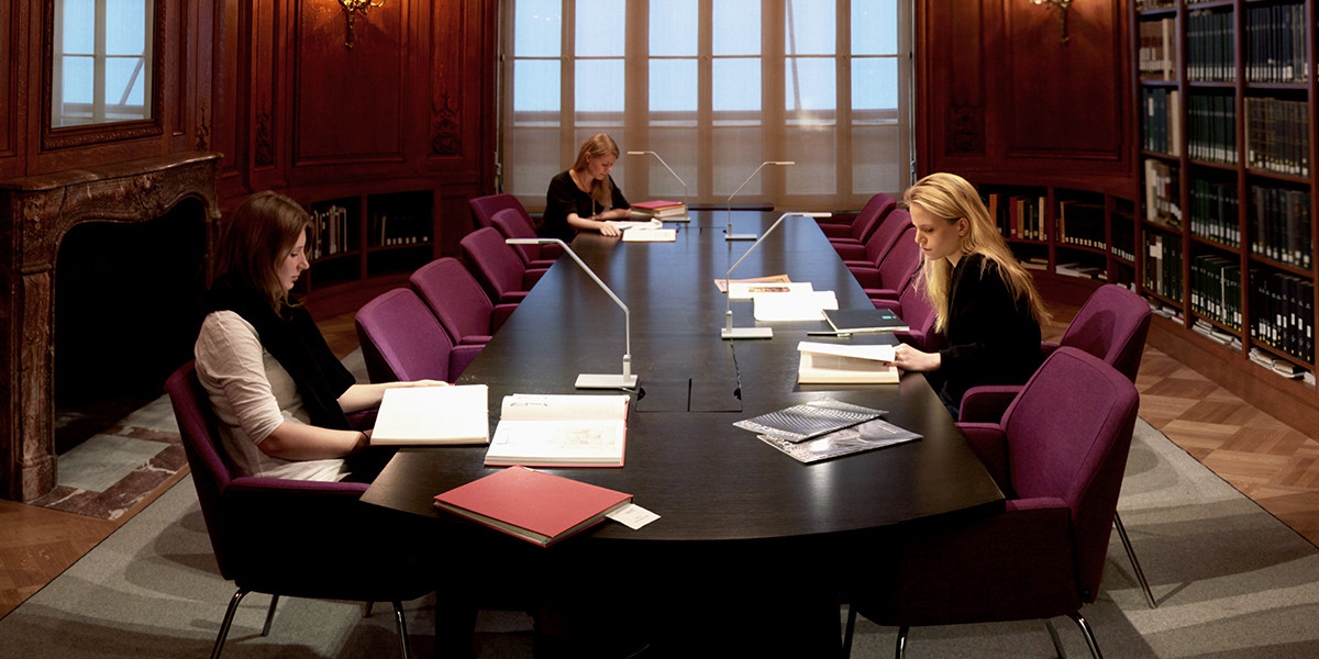 Small wood-paneled room with a large table in the center and three women seated at the table, reading. Books on shelves all around them.