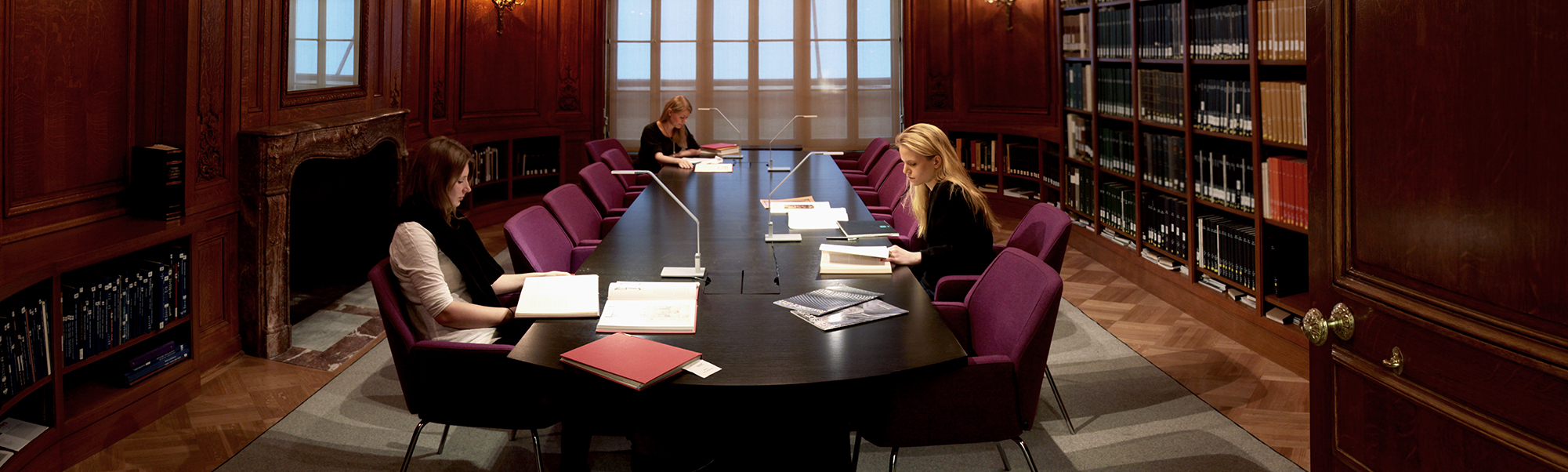 Small wood-paneled room with a large table in the center and three women seated at the table, reading. Books on shelves all around them.