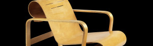 A wooden armchair, a light colored wood, with bended undulating shapes against a stark black background.