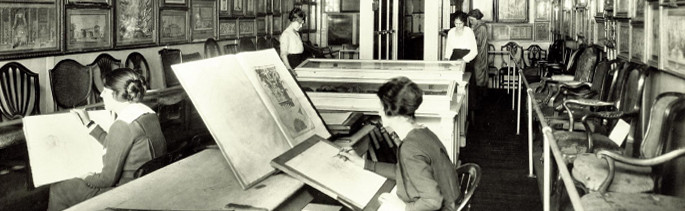 Photo of a large room. Walls covered in framed works. Women in tall leather boots with long dresses and long hair sit hunched over sketchboards, appearing to copy other works set in front of them on the table.