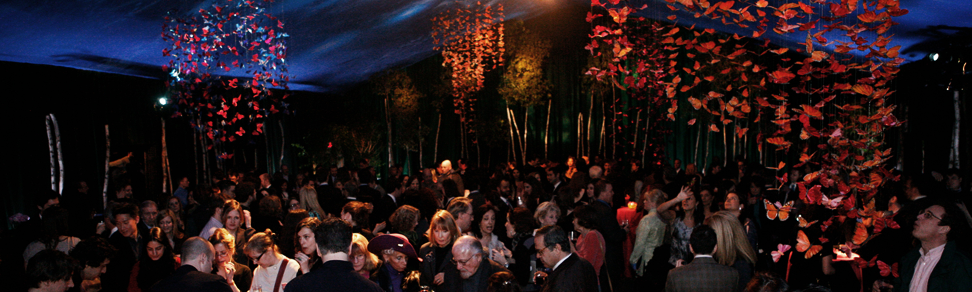 a darkened event space with dramatically lit faux butterflies draped in clumps from the ceiling, and a room full of adults in formal attire, gathering near a large bar.