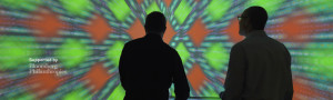 two silhouettes standing before a colorful glowing display of a repeating orange and green pattern. the pattern is blurred at the edges, as if rushing toward the camera.