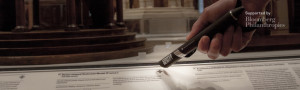 A glass case with wooden architectural models inside. a hand reaches toward the label beside the case, holding an oversized black pen, as if to touch the label with the eraser side of the pen.