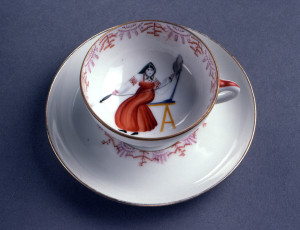 Cup with shallow bowl on raised foot, loop handle. Painted in interior of bowl with woman dressed in peasant costume, spinning with distaff; stylized border upper edge, thin gilded bands top and bottom edge of outside. Saucer round, slightly raised towards edges; painted with matching stylized border around center, two thin gilded bands bordering.