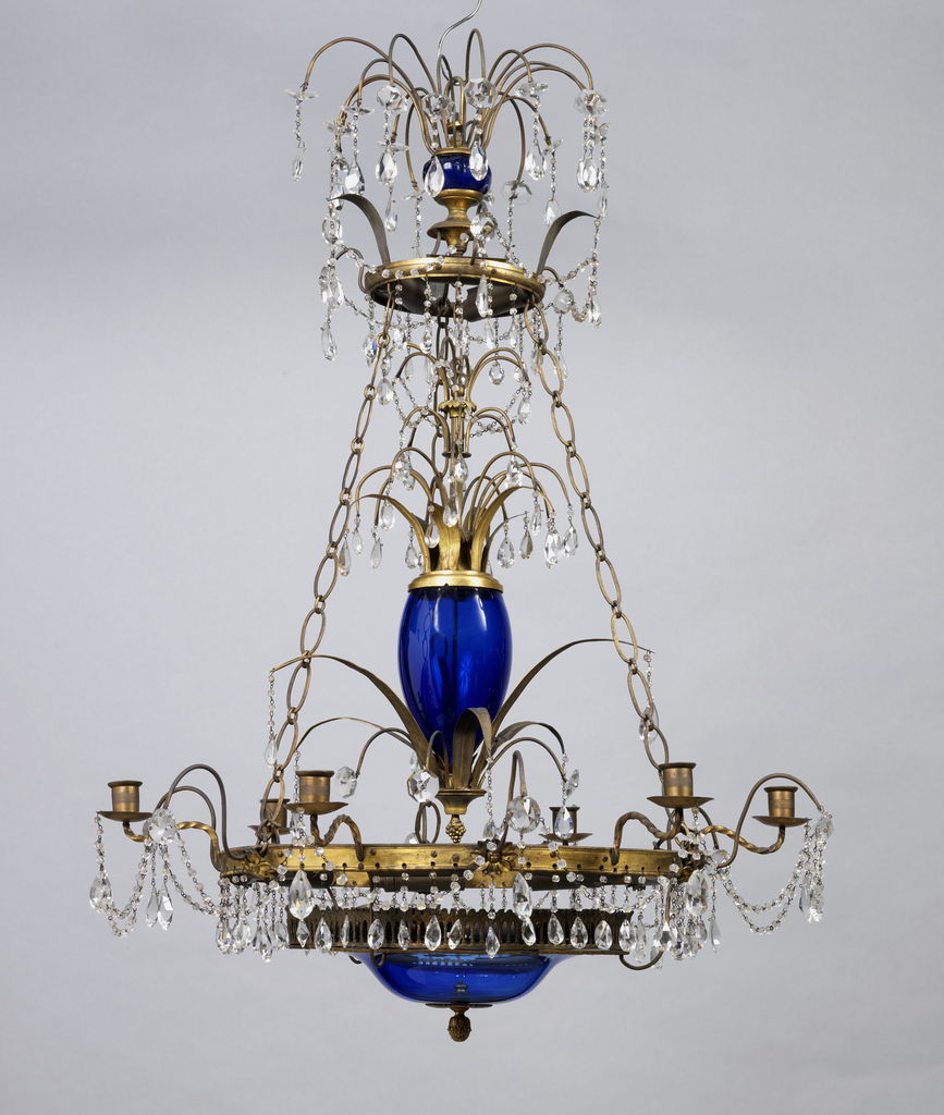 Three tiers of fountains of glass descend from the top, set off by swags of glass drops, the blown glass stems delicately engraved; gilt lower ring with six candle arms and an upper ring connected by the glass-surrounded stem and by three chains, all of metal, the lower ring supporting a blue glass disc at the base of the stem.