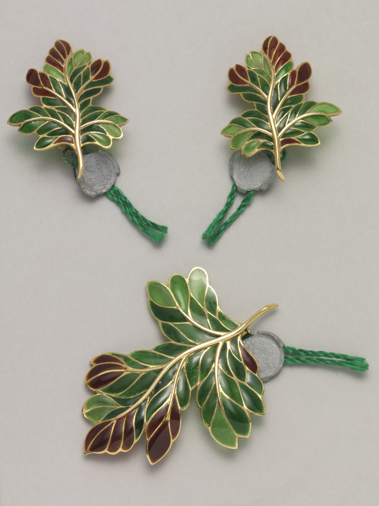 Two earclips and a brooch, each in the form of an oak leaf with a gold stem and veining, filled with deep green and dark red plique-à-jour enamel. Earclips have plastic ear cushions. Attached to each piece by a green cord is a circular stamped lead seal with mark.