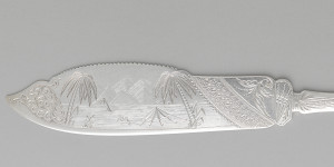 silver cake saw with engraving of palm tree landscape on the blade of the saw.