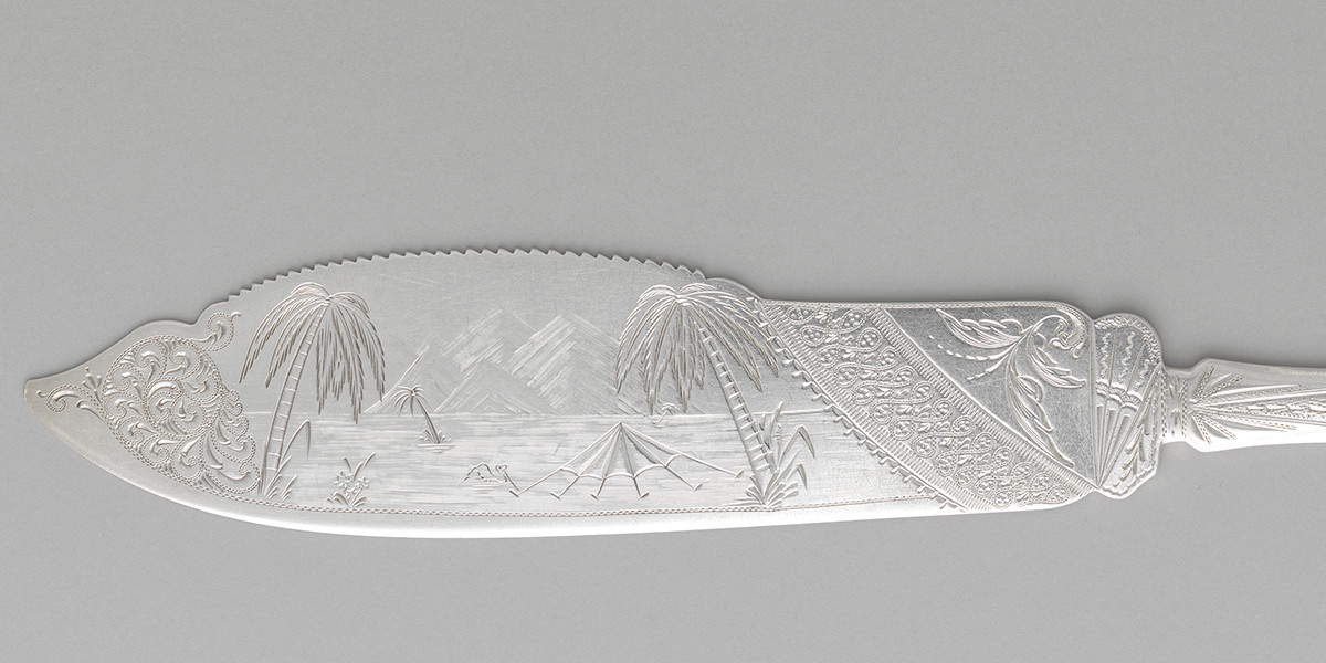silver cake saw with engraving of palm tree landscape on the blade of the saw.