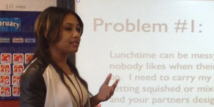 A woman stands in front of a screen, speaking and gesticulating.