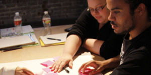 A group of four middle aged people seated around a table, touching a poster and post-it notes in the middle of the table.