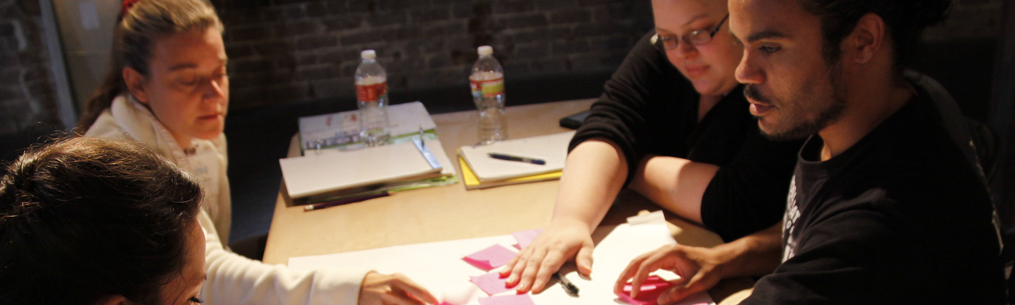 A group of four middle aged people seated around a table, touching a poster and post-it notes in the middle of the table.