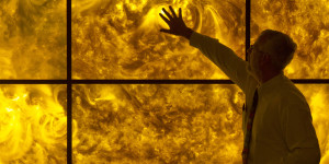 A man standing in front of six large screens displaying a single image of the firey sun. The man reaches out to touch the image.