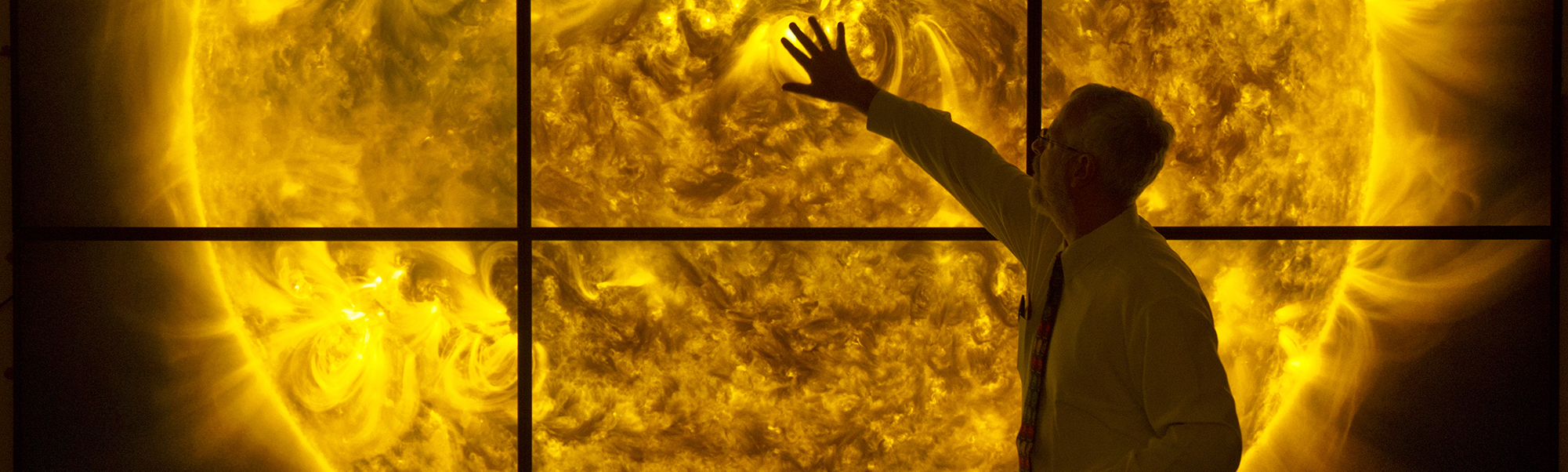 A man standing in front of six large screens displaying a single image of the firey sun. The man reaches out to touch the image.