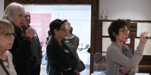 a woman in a museum setting gesticulates toward the right, with several people behind her, musing together