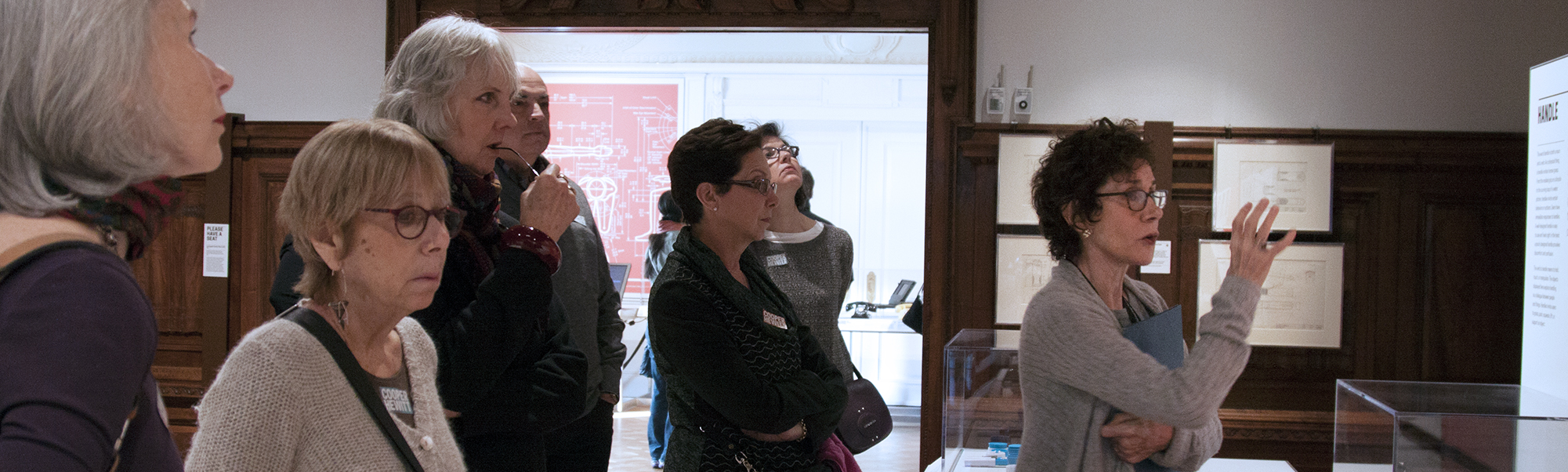 a woman in a museum setting gesticulates toward the right, with several people behind her, musing together